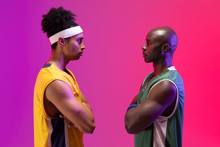Image of two diverse basketball players facing each other on purple to pink background. Sports and...
