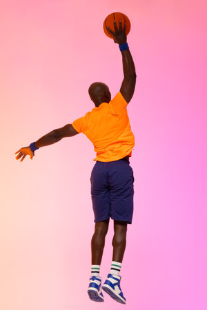 Image of african american basketball player jumping with basketball on pink to orange background....