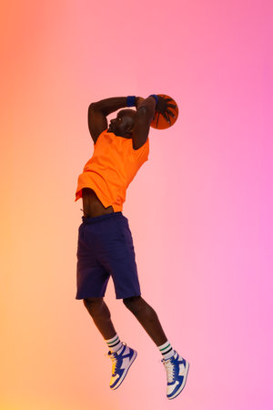 Image of african american basketball player jumping with basketball on pink to orange background. Sports and competition concept.の写真素材