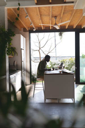 Happy african american man sitting at table in kitchen using laptop. Spending quality time at home alone.の写真素材
