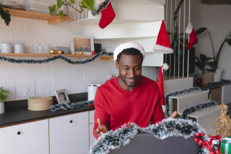 Happy african american man wearing santa hat, using laptop for video call. Spending time at home alone at christmas.の写真素材
