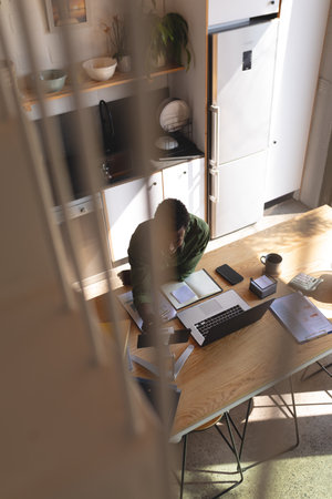 Happy african american man sitting at table in kitchen using laptop and tablet. Spending quality...