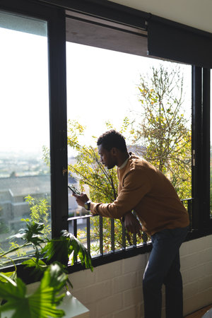 Happy african american man looking through window and using smartphone. Spending quality time at home alone.の写真素材