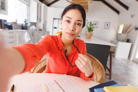 Happy biracial woman sitting at table in kitchen, having video call. Spending quality time at home...