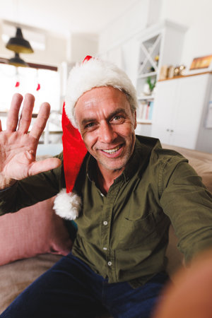 Happy caucasian man wearing santa hat, sitting on sofa in living room and having video call. Spending quality time at home alone at christmas.の写真素材