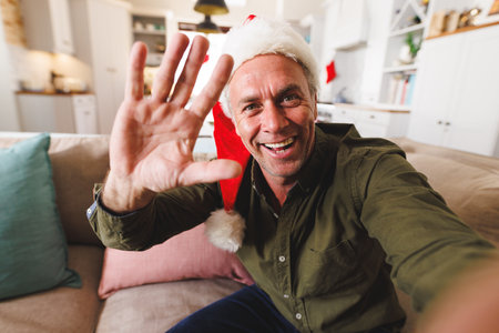 Happy caucasian man wearing santa hat, sitting on sofa in living room and having video call. Spending quality time at home alone at christmas.の写真素材