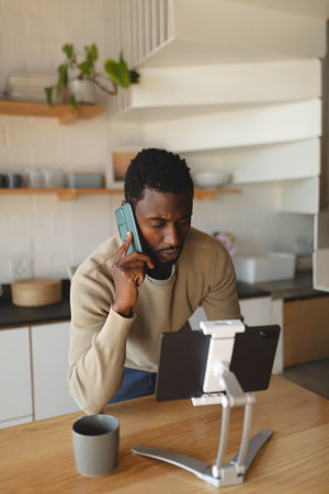 Happy african american man using tablet and talking on smartphone in kitchen. Spending quality time at home alone.の写真素材