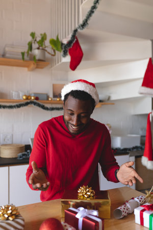 Happy african american man wearing santa hat, with christmas presents at table. Spending time at home alone at christmas.の写真素材