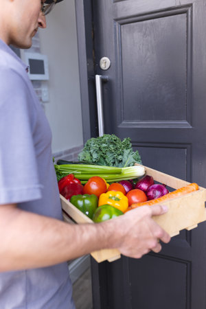 Caucasian male courier with vegetables waiting outside house door for the client. Online grocery shopping.の写真素材