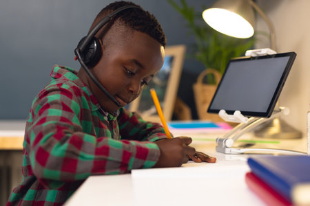 Smiling african american boy learning, using headphones and tablet with copy space at home. Home...