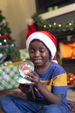 Vertical picture of african american boy holding christmas snowing ball and siting next fireplace. Christmas, childhood and celebration concept.の写真素材