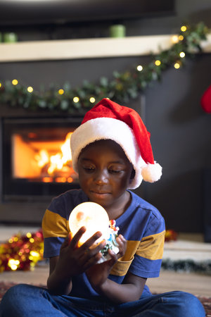 Vertical picture of african american boy holding christmas snowing ball and siting next fireplace. Christmas, childhood and celebration concept.の写真素材