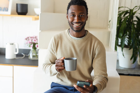 Happy african american man sitting on stairs in kitchen, drinking coffee and using smartphone. Spending quality time at home alone.の写真素材