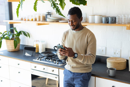 Happy african american man leaning on countertop in kitchen, using smartphone. Spending quality time at home alone.の写真素材