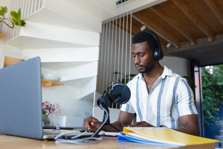 Happy african american man sitting at table in kitchen, using laptop and making vlog. Spending quality time at home alone.の写真素材