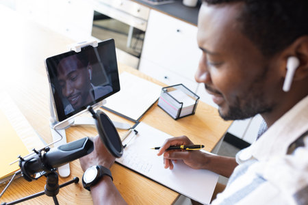Happy african american man sitting at table in kitchen, using tablet and making vlog. Spending quality time at home alone.の写真素材
