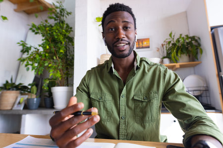 Happy african american man sitting at table in kitchen, having video call. Spending quality time at...