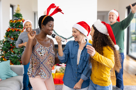 Image of happy diverse female friends singing karaoke celebrating christmas at home. Christmas, celebration, tradition, friendship, inclusivity and lifestyle concept.の写真素材