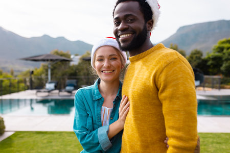 Portrait of happy diverse couple in santa hats to celebrate christmas smiling outdoors. Christmas, celebration, tradition, friendship, inclusivity and lifestyle concept.の写真素材