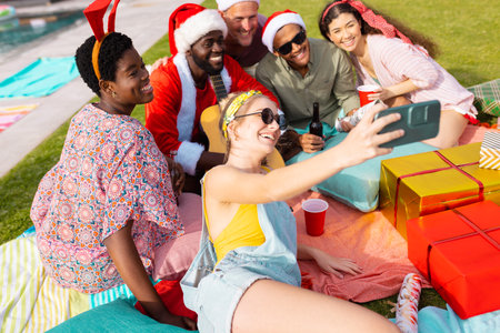 Happy diverse friends with santa hats having picnic and taking selfie in garden at christmas. Christmas, celebration, friendship, inclusivity and lifestyle concept.の写真素材