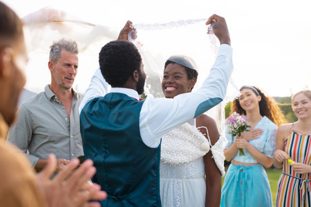 Happy african american couple holding veil and smiling during wedding. Wedding day, friendship, inclusivity and lifestyle concept.の写真素材