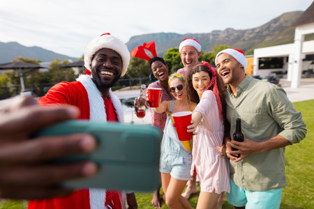 Happy diverse friends with santa hats taking selfie in garden at christmas. Christmas, celebration, friendship, inclusivity and lifestyle concept.の写真素材