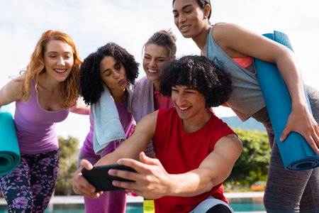 Diverse friends using smartphone and holding yoga mats in garden. Health, celebration, friendship,...