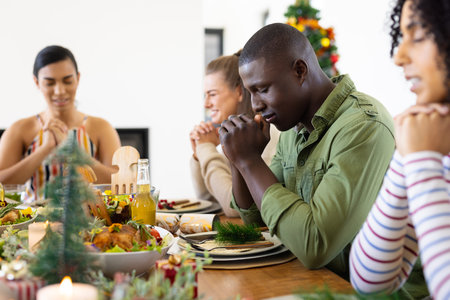 Happy diverse friends sitting at table and praying before dinner at christmas. Christmas, celebration, friendship, inclusivity and lifestyle concept.の写真素材