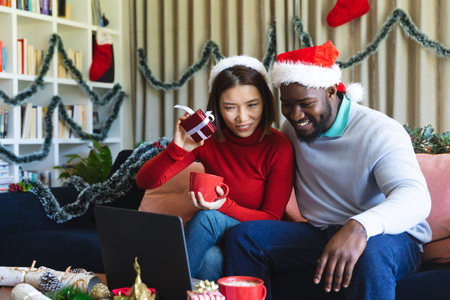 Happy diverse couple wearing santa claus hats, using laptop for video call. Spending quality time at christmas together.の写真素材