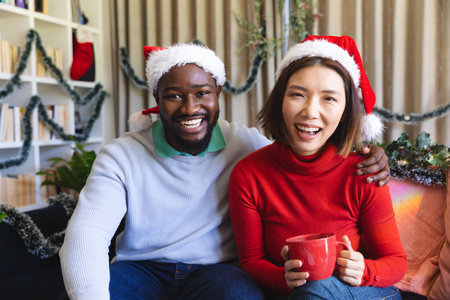Portrait of happy diverse couple wearing santa claus hats looking at camera and smiling. Spending quality time at christmas together.の写真素材