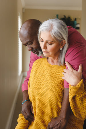 Vertical image of thoughtful, sad diverse senior couple embracing and looking down at home....