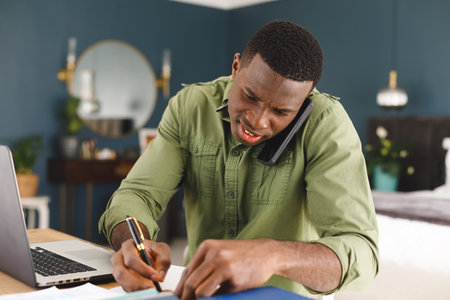 Smiling african american man working at home in bedroom using laptop, talking on phone and writing. Communication, working from home, domestic life and inclusivity concept.の写真素材