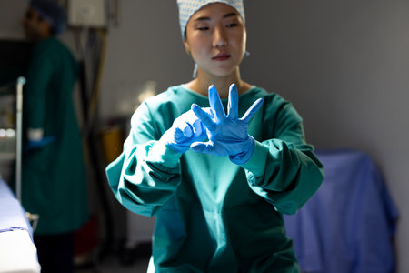 Asian female surgeon in surgical cap and gown putting on gloves in operating theatre. Hospital, medical and healthcare services.の写真素材