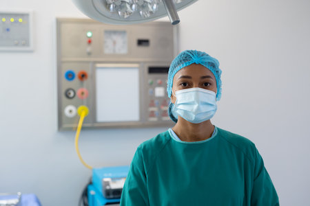 Portrait of biracial female healthcare worker in surgical cap, mask and gown in operating theatre. Hospital, medical and healthcare services.の写真素材