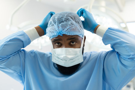 African american male surgeon in gown and cap tying on mask in operating theatre. Hospital, medical...