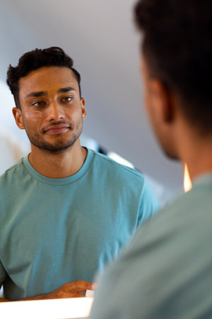 Smiling biracial man looking at himself in bathroom mirror after applying face cream. Lifestyle, self care and domestic life, unaltered.の写真素材