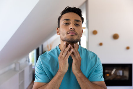 Biracial man touching his neck looking in bathroom mirror. Lifestyle, self care and domestic life, unaltered.の写真素材