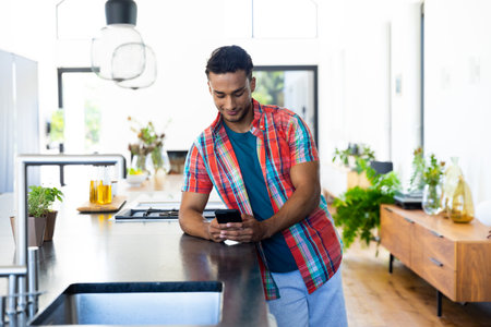 Happy biracial man leaning on countertop and using smartphone in kitchen. Lifestyle, technology, communication and domestic life, unaltered.の写真素材