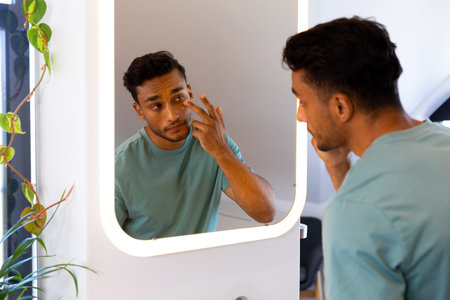 Biracial man looking at himself in bathroom mirror and applying under eye cream. Lifestyle, self care and domestic life, unaltered.の写真素材