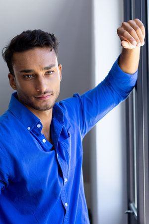Portrait of biracial man leaning on window and looking at camera. Relaxation, lifestyle and domestic life, unaltered.の写真素材