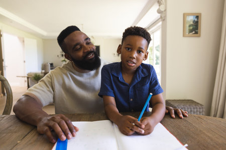 Happy african american father and son sitting at table with notebook and having video call. Communication, video calls, education, family and domestic life.の写真素材