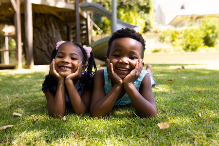Portrait of cute african american siblings with hands on chins lying on grassy field in park. Unaltered, family, togetherness, childhood, smiling, enjoyment and weekend concept.の写真素材