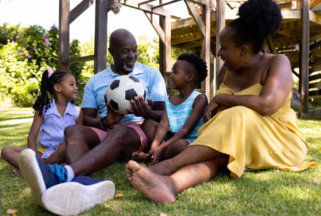 African american grandparents and grandchildren with soccer ball sitting on grassy land in park. Unaltered, family, togetherness, childhood, sports, retired, ball, enjoyment and weekend concept.の写真素材