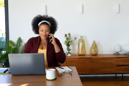 Happy plus size african american woman sitting at table, using laptop and talking on smartphone. Communication, working from home, body inclusivity and lifestyle, unaltered.の写真素材
