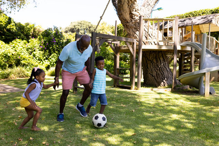 African american grandfather playing soccer with cheerful grandchildren in park. Unaltered, family,...