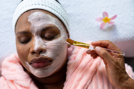 Relaxed plus size african american woman having beauty face mask applied to face at spa. Self care, health and beauty, body inclusivity and lifestyle, unaltered.の写真素材