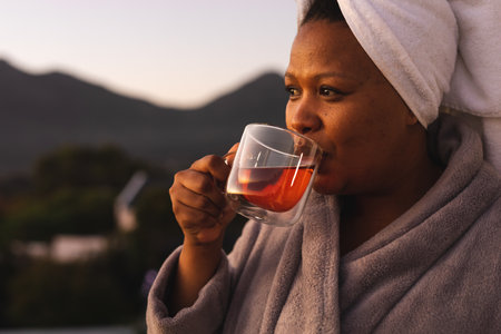 Happy plus size african american woman wearing robe and towel on head, drinking tea on balcony. Relaxation, body inclusivity, lifestyle and self care, unaltered.の写真素材
