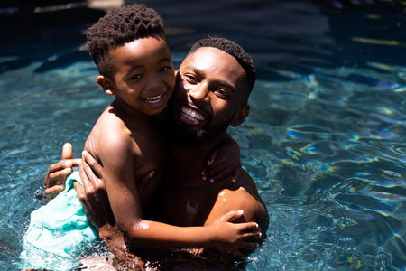Portrait of cheerful african american shirtless father carrying son while swimming in pool at...