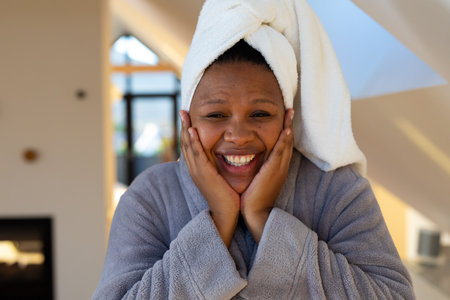 Happy plus size african american woman wearing robe, holding her face and smiling in bathroom. Self care, health and beauty, body inclusivity and lifestyle, unaltered.の写真素材