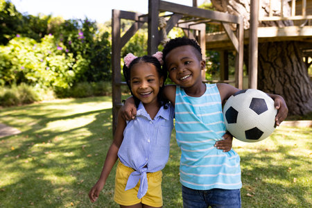 Portrait of smiling african american siblings with soccer ball standing in park. Unaltered, family, togetherness, childhood, sports, enjoyment and weekend concept.の写真素材